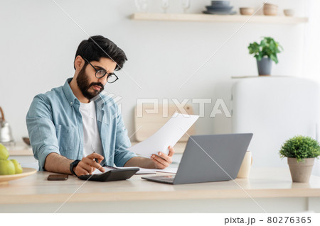 Paying bills, taxes at home online. Young arab man using calculator and laptop computer, sitting at kitchen, copy space 80276365