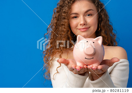 Curly-haired young woman holding piggy bank against blue background 80281119