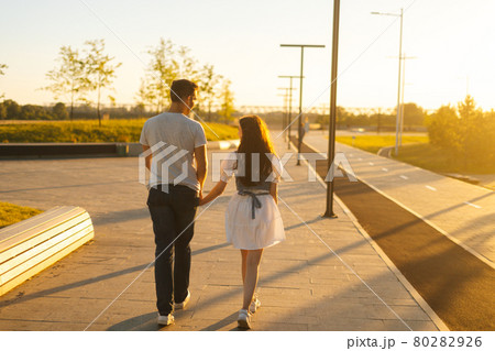 Rear view of young couple in love holding hands walking in city park in sunny summer day on background of bright sunlight. Rear view of young couple in love holding hands walking in city park in sunny summer day on background of bright sunlight. 80282926