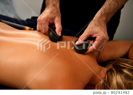 Masseur placing therapeutic hot stones on woman's back and doing Ayurvedic hot stone massage in modern spa salon. Body care concept Masseur placing therapeutic hot stones on woman's back and doing Ayurvedic hot stone massage in modern spa salon. Body care concept 80291482