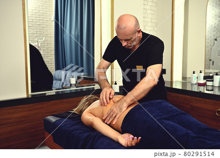 Professional masseur massaging woman's back in a wellness clinic. A mature chiropractor gives a therapeutic massage to a client of the SPA center. Professional masseur massaging woman's back in a wellness clinic. A mature chiropractor gives a therapeutic massage to a client of the SPA center. 80291514