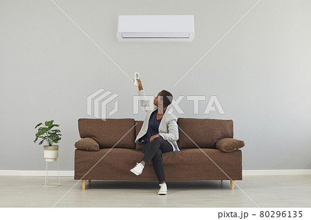 Black woman sitting on couch and switching her air conditioner with remote control 80296135