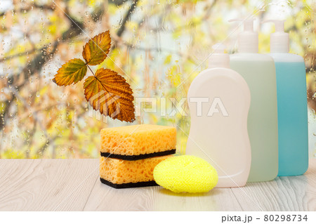 Bottles of dishwashing liquid and sponges in front of the window with water drops. 80298734