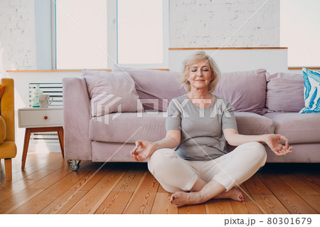 Senior adult smiling woman practicing yoga at home living room. Elderly relaxed female sitting in lotus pose and meditating zen like. Senior adult smiling woman practicing yoga at home living room. Elderly relaxed female sitting in lotus pose and meditating zen like. 80301679