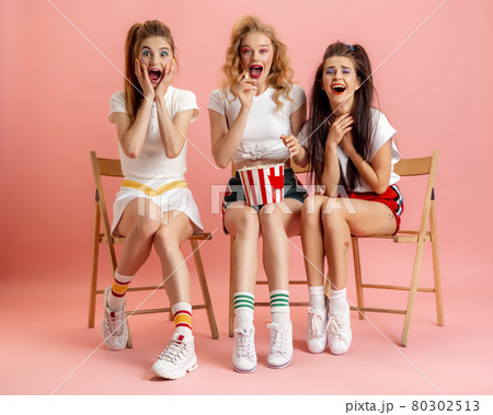 Three young women in retro 90s fashion style, outfits watching movie isolated over pink studio background. Concept of eras comparison, beauty, fashion and youth. 80302513