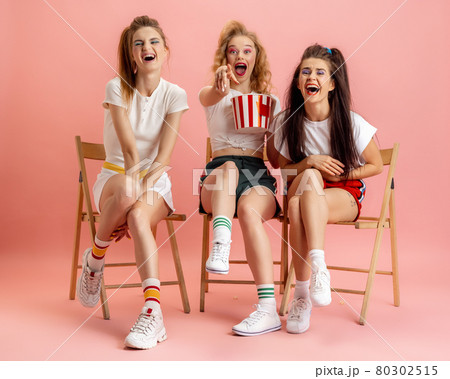 Three young women in retro 90s fashion style, outfits watching movie isolated over pink studio background. Concept of eras comparison, beauty, fashion and youth. 80302515