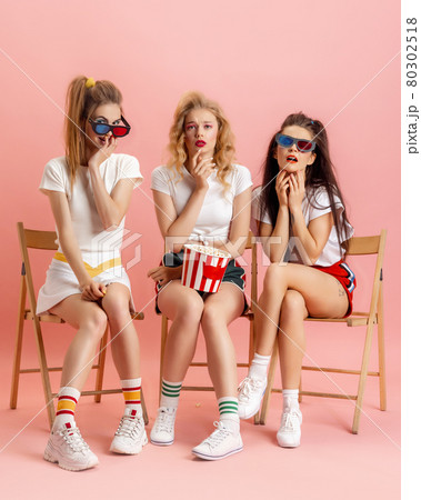 Three young women in retro 90s fashion style, outfits watching movie isolated over pink studio background. Concept of eras comparison, beauty, fashion and youth. 80302518