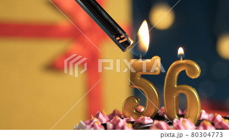 Birthday cake number 56 golden candles burning by lighter, background gift yellow box tied up with red ribbon. Close-up Birthday cake number 56 golden candles burning by lighter, background gift yellow box tied up with red ribbon. Close-up 80304773