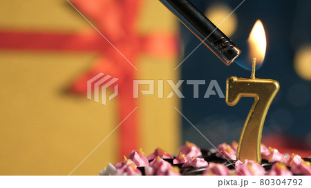 Birthday cake number 7 golden candles burning by lighter, background gift yellow box tied up with red ribbon. Close-up Birthday cake number 7 golden candles burning by lighter, background gift yellow box tied up with red ribbon. Close-up 80304792