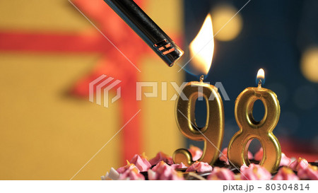 Birthday cake number 98 golden candles burning by lighter, background gift yellow box tied up with red ribbon. Close-up 80304814