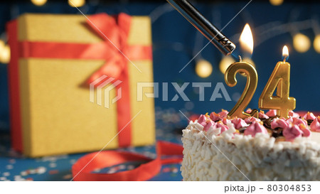 White birthday cake number 24 golden candles burning by lighter, blue background with lights and gift yellow box tied up with red ribbon. Close-up 80304853
