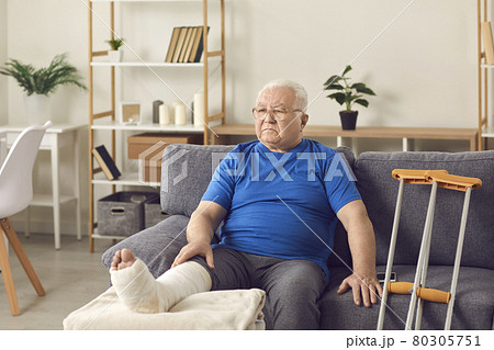 Disappointed senior mature man sitting on sofa with broken leg in cast 80305751