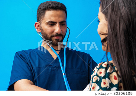 Arab doctor in a blue uniform standing with the stethoscope and listening to the heartbeat of a woman patient Arab doctor in a blue uniform standing with the stethoscope and listening to the heartbeat of a woman patient 80306109