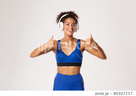 Healthy athletic african-american woman in blue sportswear, showing thumbs-up, listening music in headphones during exercises, workout over white background 80307257