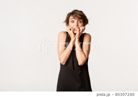 Image of scared and insecure brunette woman in black dress, trembling from fear and biting fingernails, standing over white background 80307280