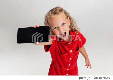 Showing device screen. One little screaming preschool girl holding smartphone isolated over white studio background. Showing device screen. One little screaming preschool girl holding smartphone isolated over white studio background. 80307685