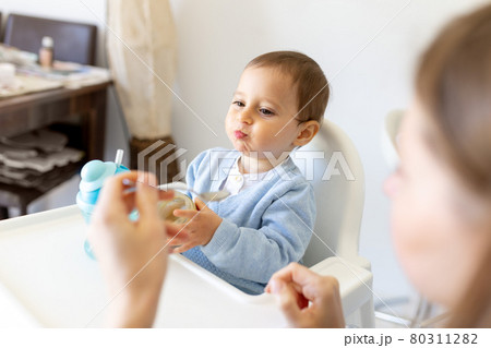 Mother feed little toddler baby boy with full mouth baby food, from plastic spoon in high chair view from back. Mother feed little toddler baby boy with full mouth baby food, from plastic spoon in high chair view from back. 80311282