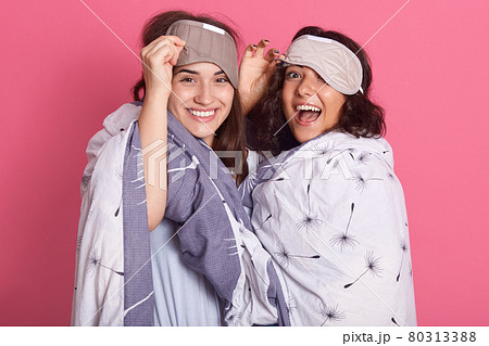 Two beautiful attractive friends laughing sincerely, having blindfold over foreheads, wearing pajamas, hugging, spending time together, ladies wrapped in blanket. Two beautiful attractive friends laughing sincerely, having blindfold over foreheads, wearing pajamas, hugging, spending time together, ladies wrapped in blanket. 80313388