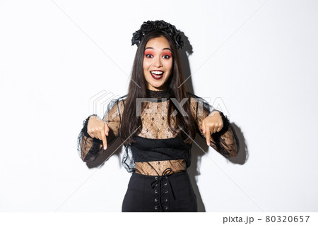 Happy beautiful asian woman in halloween gothic dress, pointing fingers down as showing announcement, demonstrating your promo, standing over white background Happy beautiful asian woman in halloween gothic dress, pointing fingers down as showing announcement, demonstrating your promo, standing over white background 80320657