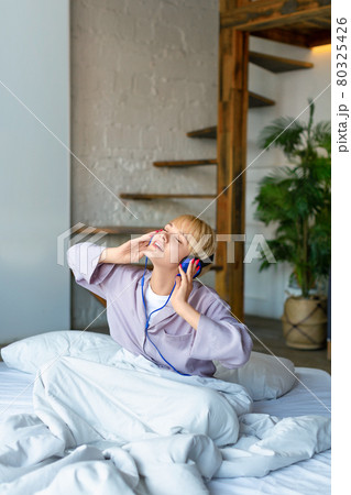 A woman woke up and listens to music through headphones while sitting in bed. She sings along while listening to her favorite song. A woman woke up and listens to music through headphones while sitting in bed. She sings along while listening to her favorite song. 80325426