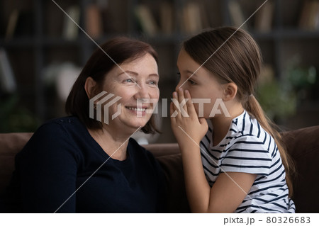 Girl telling secret to middle aged 60s grandma, whispering in ear 80326683