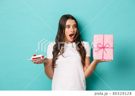 Young excited woman celebrating her birthday, holding bday cake and gift, standing amazed over blue background Young excited woman celebrating her birthday, holding bday cake and gift, standing amazed over blue background 80329128