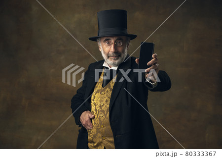 Cute senior man, gentleman, aristocrat or actor talking on phone isolated on dark vintage background. Retro style, comparison of eras concept. 80333367