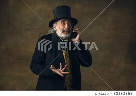 Cute senior man, gentleman, aristocrat or actor talking on phone isolated on dark vintage background. Retro style, comparison of eras concept. 80333368
