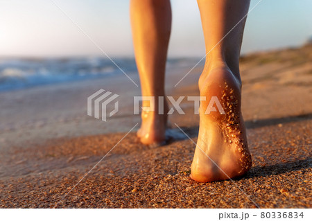 Close up detail bottom behind pov view of pretty female person barefoot legs heel walking by scenic sandy ocean or sea beach at warm golden sunrise morning. Wild nature travel and vacation concept 80336834