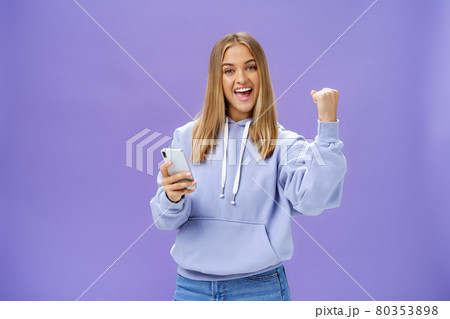 Yes we did it. Pleased excited woman in hoodie raising fist in celebration and success gesture smiling broadly triumphing holding smartphone reacting to positive news in cellphone over purple wall 80353898