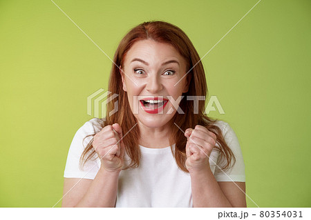 Close-up lucky enthusiastic cute redhead joyful middle-aged woman pump fists vigorous excitement celebratory smiling broadly winning celebrating triumphing success good news green background 80354031