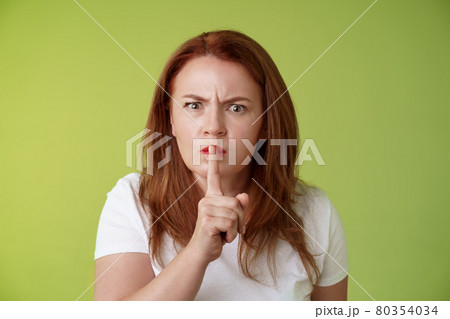 Not speak during exam. Strict serious-looking displeased middle-aged redhead woman frowning disappointed hushing say shush index finger pressed lips keep quiet gesture green background 80354034