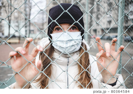 The girl in the medical mask is in the street in isolation and is holding fence. 80364584