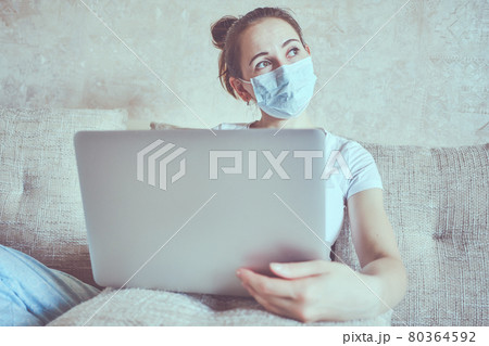 A girl in a medical mask is in quarantine at home holding a laptop and look out the window. 80364592