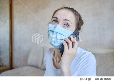 The girl in a medical mask is on self-isolation at home and speaks on the phone with relatives. 80364606