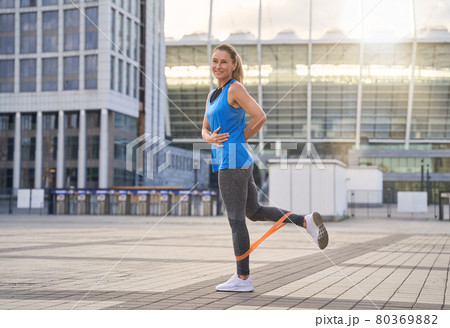 Full length shot of smiling sporty woman in sportswear using resistance band while standing outdoors, exercising in the city Full length shot of smiling sporty woman in sportswear using resistance band while standing outdoors, exercising in the city 80369882
