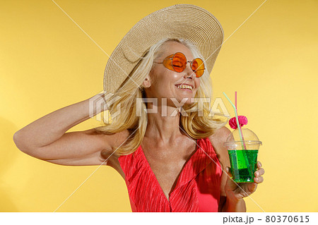 Relaxed blonde woman in sunglasses, red swimsuit and straw hat smiling with eyes closed, adjusting her hat and holding cocktail drink while posing isolated on yellow background Relaxed blonde woman in sunglasses, red swimsuit and straw hat smiling with eyes closed, adjusting her hat and holding cocktail drink while posing isolated on yellow background 80370615