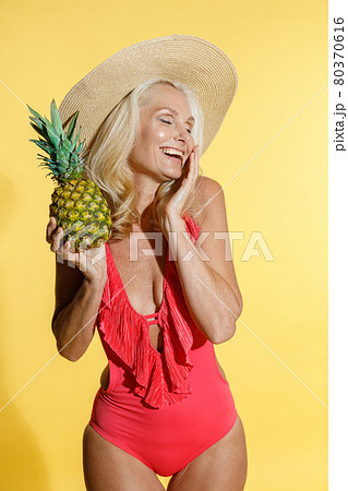 Careless mature blonde woman in red swimsuit and straw hat laughing with eyes closed, holding fresh pineapple fruit while posing isolated on yellow background in studio Careless mature blonde woman in red swimsuit and straw hat laughing with eyes closed, holding fresh pineapple fruit while posing isolated on yellow background in studio 80370616