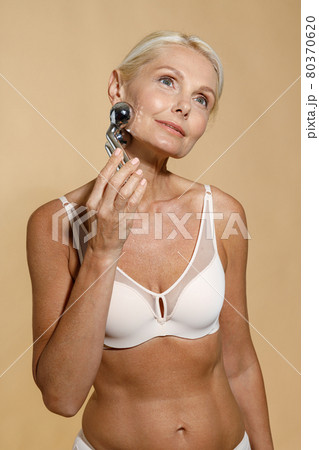 Beautiful mature blonde woman in underwear with glowing skin using metal face roller, looking away, posing isolated over beige background 80370620