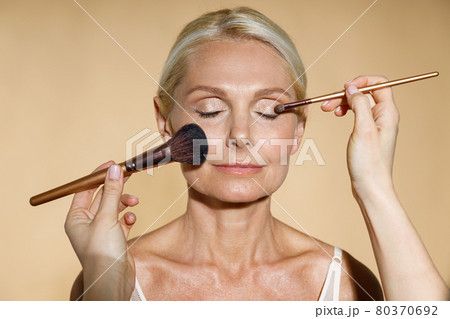 Close up of hands of skillful artist doing natural makeup for mature blonde woman isolated over beige background. The woman closed her eyes with serenity Close up of hands of skillful artist doing natural makeup for mature blonde woman isolated over beige background. The woman closed her eyes with serenity 80370692