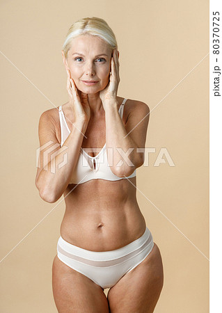 Woman in underwear with fit body looking at camera and touching facial skin, standing isolated over beige background 80370725