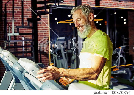 Side view of a happy middle aged athletic man in sportswear adjusting speed on a treadmill while having cardio workout in a gym or sport club 80370856