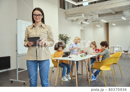 Beautiful young female teacher in casual wear smiling at camera and holding tablet pc while standing in a classroom. Kids working on diy robot in the background 80370936