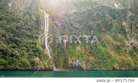 Huge waterfall falling from green cliffs with two small canoes at the bottom for scale. Location is Stirling Falls in Milford Sound, Fiordland NP 80375704