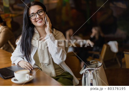 Pretty young business woman talking smartphone sitting at table in cafe with laptop and coffee cup 80376195