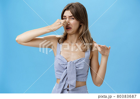 Gross smells awful. Portrait of displeased funny young slim woman with tattoo squeezing nose with two fingers not sniff disfusting reek crying from stink standing intense over blue background Gross smells awful. Portrait of displeased funny young slim woman with tattoo squeezing nose with two fingers not sniff disfusting reek crying from stink standing intense over blue background 80383687