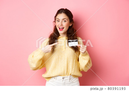 Portrait of young brunette caucasian girl pointing finger at plastic credit card, recommending bank, going on shopping, standing over pink background 80391193