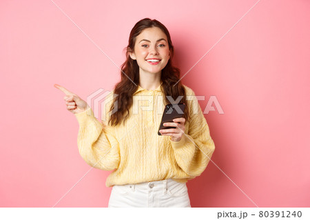 Beautiful modern girl holding smartphone and pointing finger aside, showing promotional text on copyspace while using mobile phone, standing over pink background 80391240