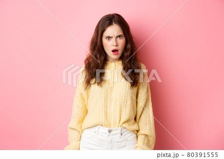 Shocked and confused brunette woman gasping, frowning and looking offended at camera, feeling insulted or displeased, standing over pink background 80391555