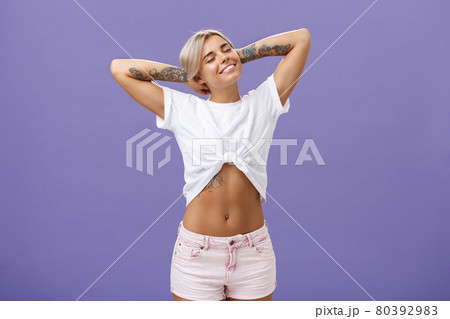 Great feelings after sunbathing. Chill and relaxed attractive young female in cropped top with perfect body and tattoos holding hands behind head, stretching with delight and smiling with closed eyes 80392983